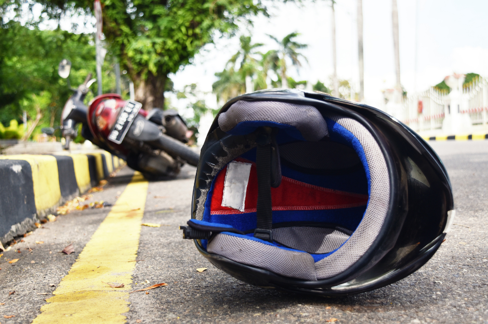Fallen black motorcycle helmet lies on a sunlit road beside a yellow-and-black curb, with a blurred motorbike behind it, suggesting a crash.