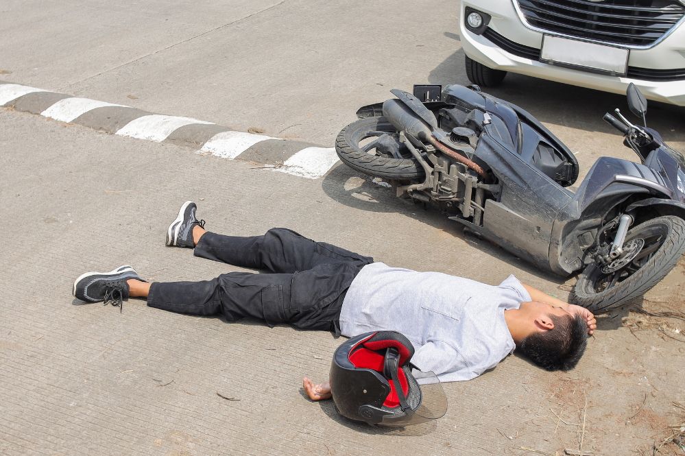 Traffic accident scene with an injured man lying on pavement beside a fallen scooter, helmet nearby, and a car stopped behind him.