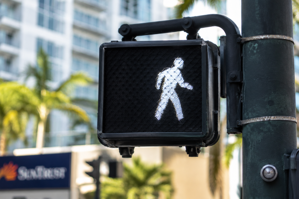 Illuminated white walking figure on a black pedestrian crossing signal, mounted to a street pole against palm trees and city buildings.