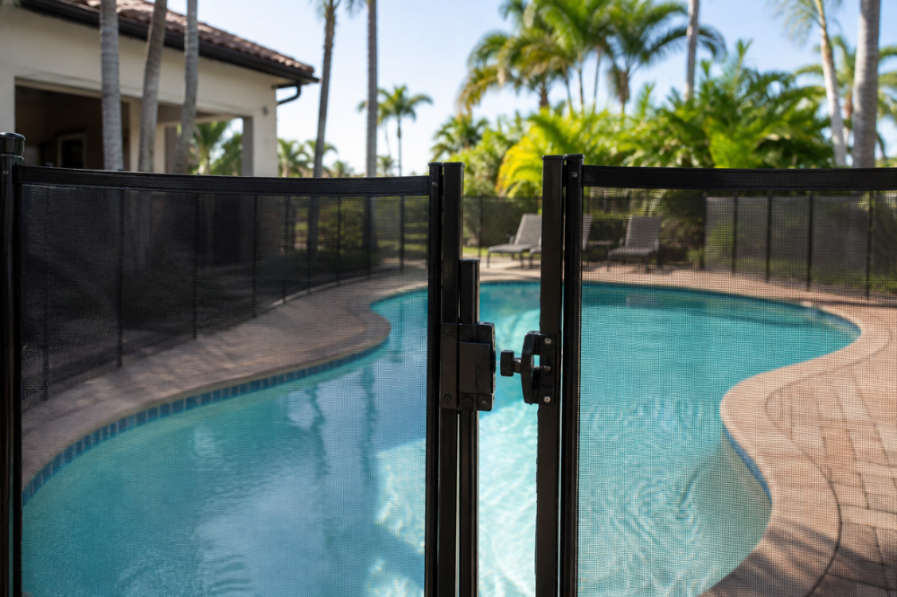 Black mesh pool safety gate opens onto a turquoise swimming pool, tiled deck, palm trees, and a sunny tropical home beyond.