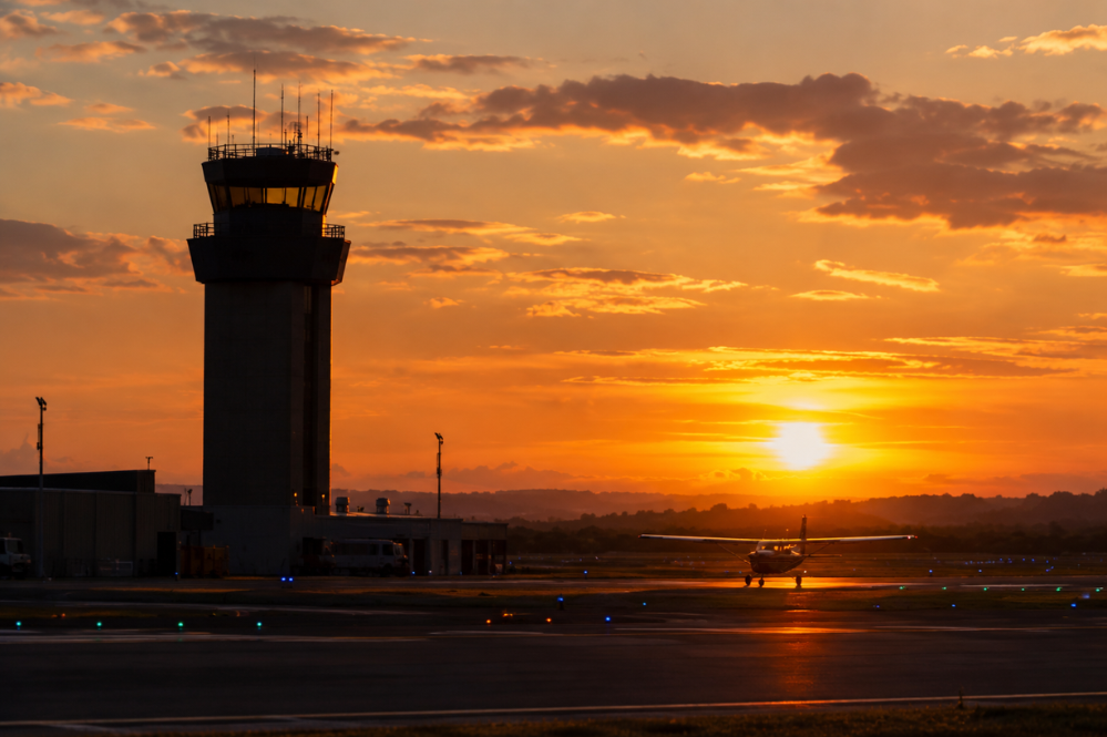 Airport control tower and small aircraft silhouetted against a vivid orange sunset, with glowing runway lights creating a calm, dramatic mood.