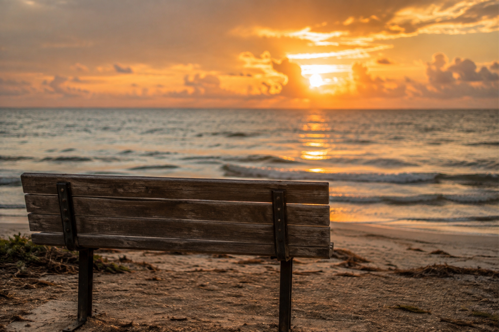 Empty wooden bench faces a calm ocean at sunset, with golden light reflecting across the waves and a peaceful, contemplative mood.